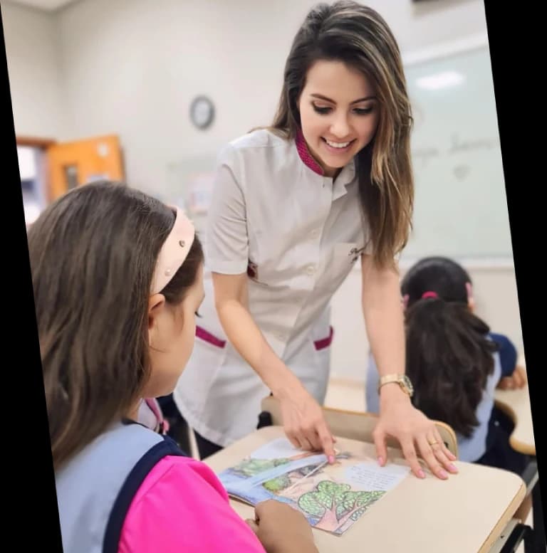 Tia Pri em sala de aula, debruçada sobre a mesa ajudando uma aluna com a atividade